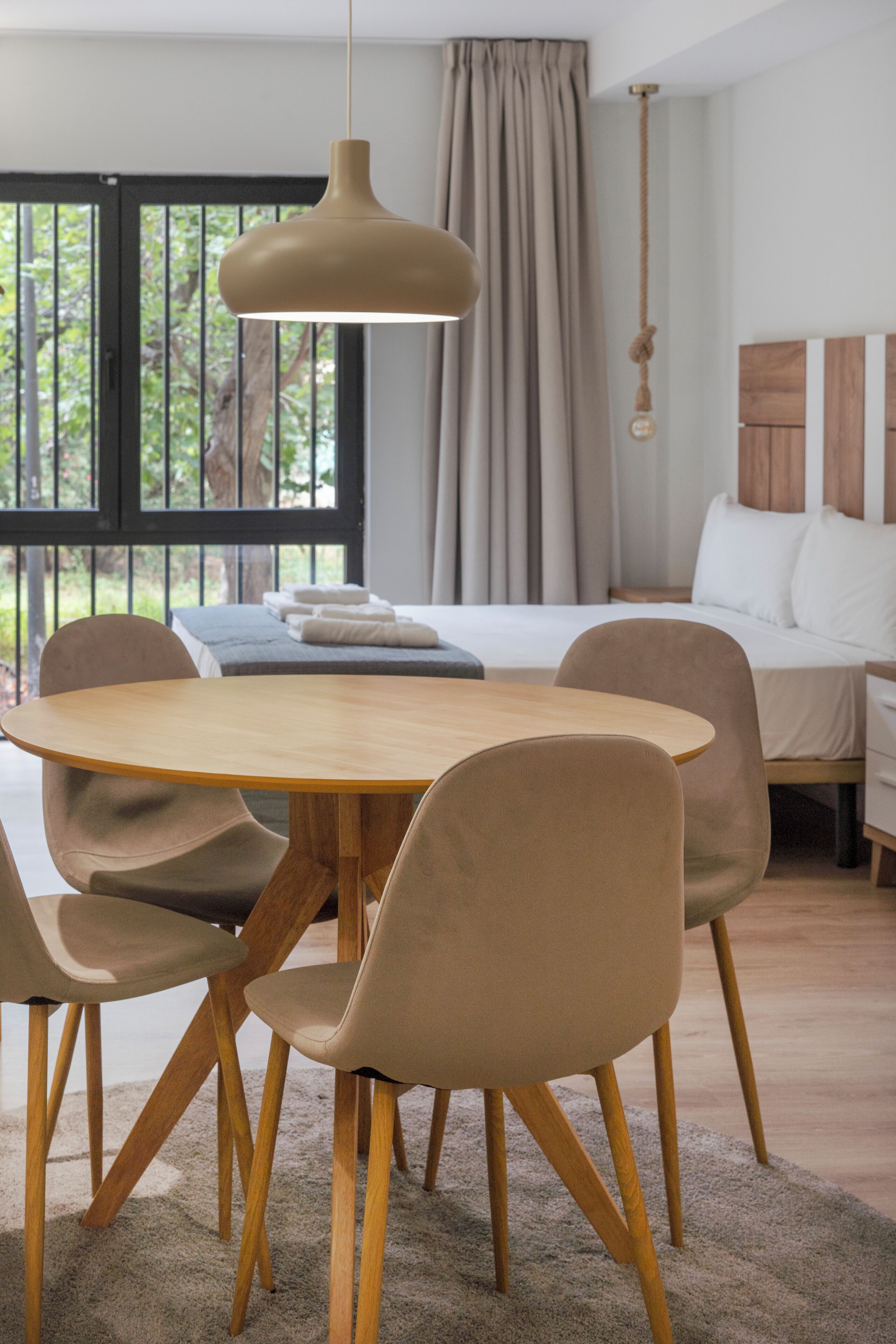 Un espace repas moderne avec une table ronde en bois et quatre chaises, un luminaire suspendu et une vue sur une chambre à coucher avec un lit et des rideaux.