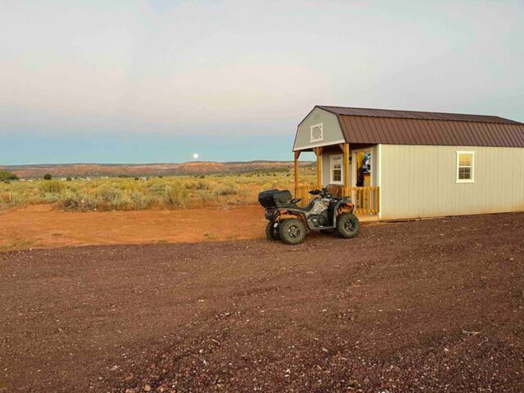 Um pequeno edifício com telhado de metal e uma porta, localizado em área rural com chão de terra e vegetação escassa.