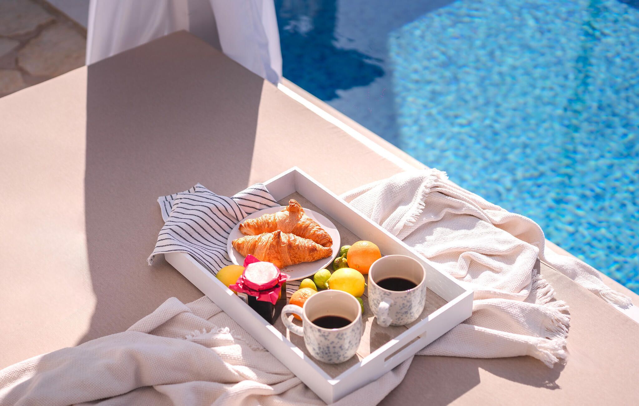 A tray with croissants, fruits, and two cups of coffee on a lounge chair by a pool.