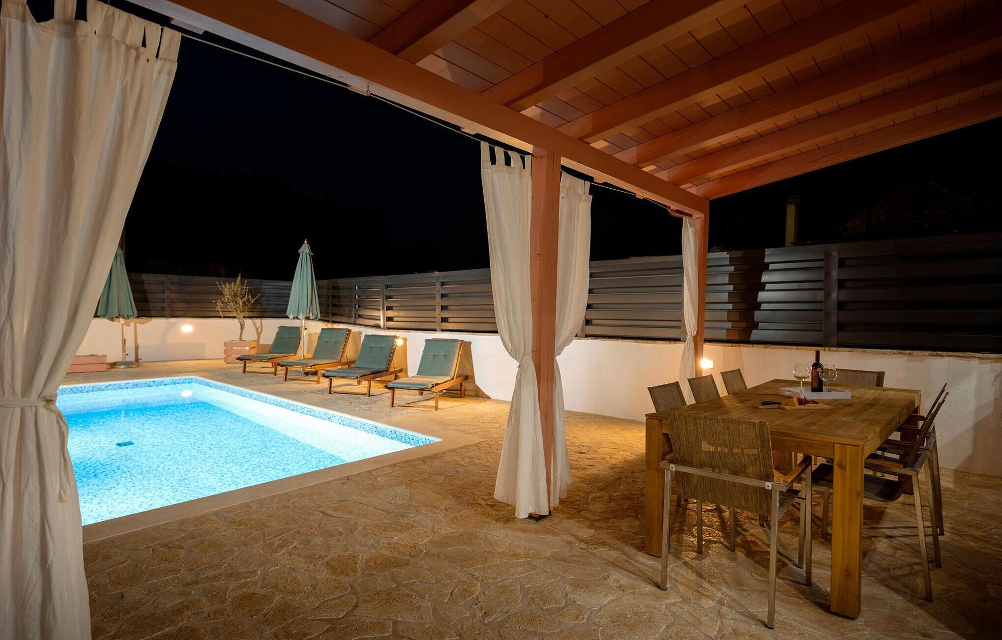 An outdoor pool area with a wooden pergola, lounge chairs, and a dining table with chairs.