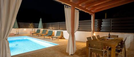 An outdoor pool area with a wooden pergola, lounge chairs, and a dining table with chairs.
