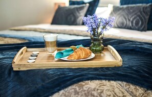 Ein Holztablett mit einem Croissant, einer Tasse Kaffee und einer Blumenvase auf einem Bett mit einer dunkelblauen Decke.