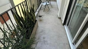 Un balcone stretto con tavolo e sedie bianche, una pianta in vaso e un parapetto in metallo.