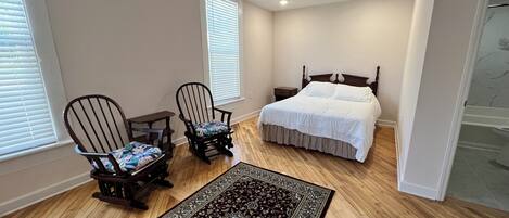 A bedroom with a wooden floor, a bed with white bedding, two rocking chairs, a nightstand, and a large patterned rug.