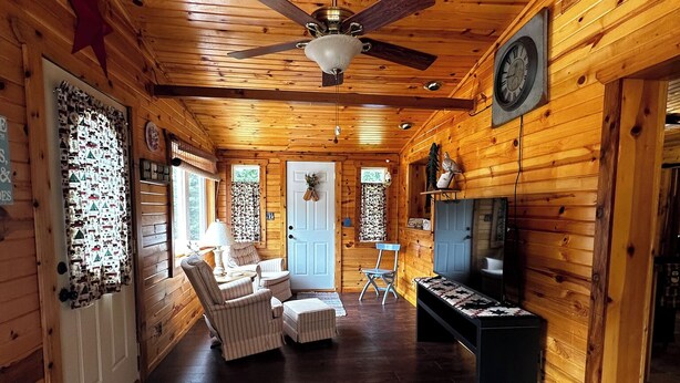 A cozy cabin interior with a ceiling fan, wooden walls, a fireplace, and a seating area with a sofa and chairs. - Cute cabin on Big Pine Lake with a large fenced yard and lake access (Finlayson)