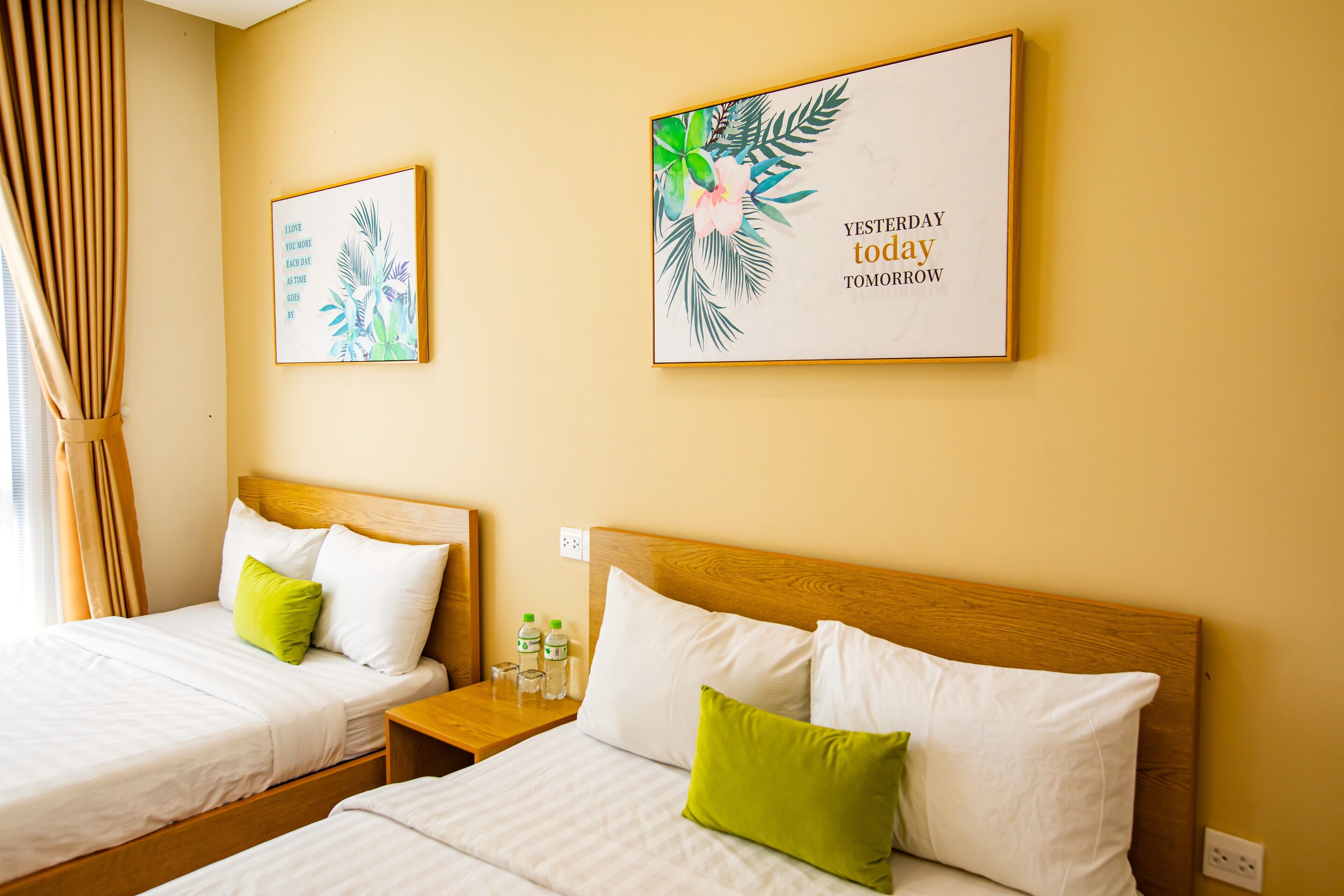 Two single beds with wooden headboards, white bedding, and green pillows. There are two framed pictures on the wall, one with a tropical design and text, and another with a similar theme.