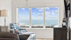 Una habitación con un gran ventanal con vista al océano, un ventilador de techo, una lámpara y un televisor sobre un mueble.