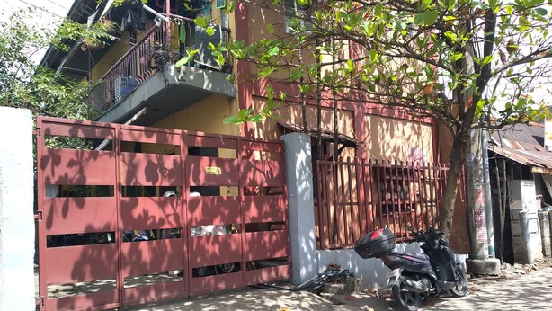 Portilla varustettu asuinalue, jossa on punainen portti, sen lähellä pysäköity moottoripyörä ja puu, jonka varjo ulottuu maahan.