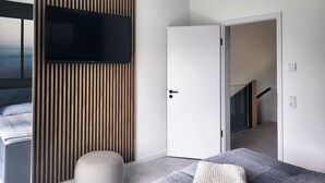 Moderni hotellihuone, jossa on suuri televisio, joka on kiinnitetty puiseen seinäpaneeliin, valkoinen ovi ja näkymä portaikkoon.