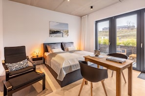 A bedroom with a bed, a desk, a chair, a nightstand, and a window with a view.