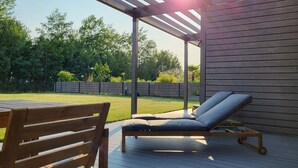 Une terrasse en bois avec un fauteuil de relaxation et une table, une pergola offrant une vue sur le jardin, et un mur en bois.