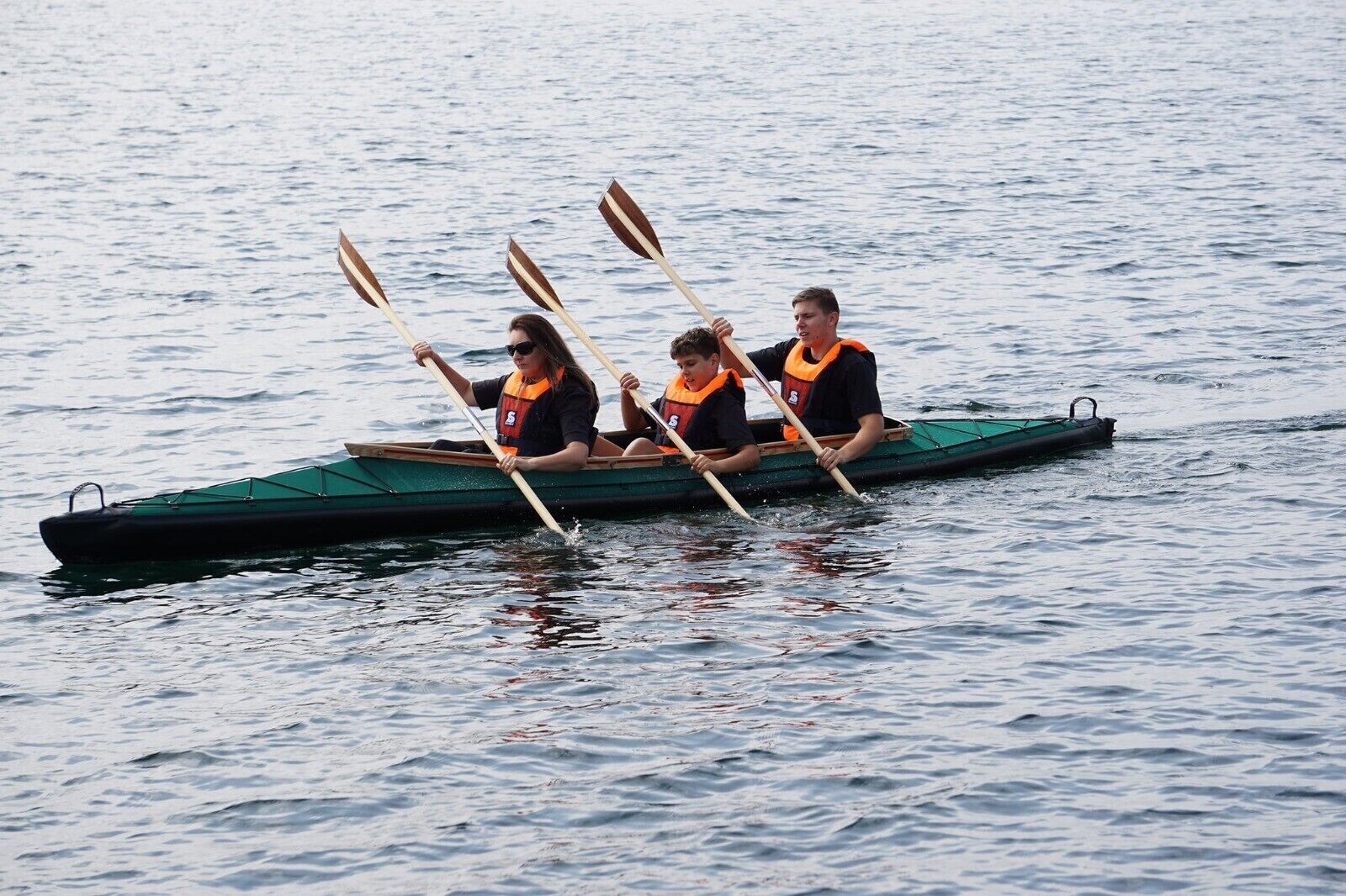 A rowing boat with three people wearing life jackets, paddling on calm water.