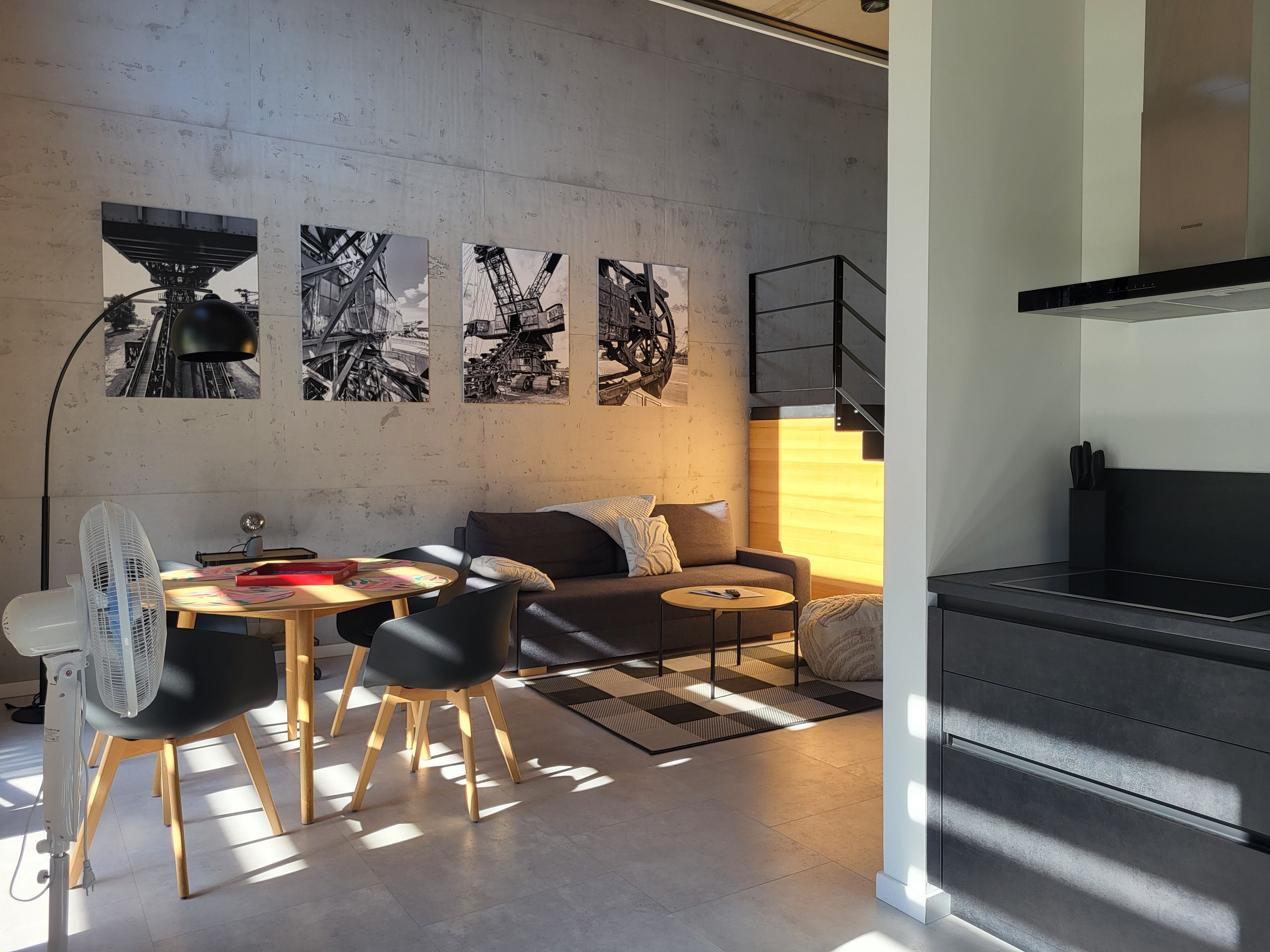 A modern living space with a sofa, dining table, chairs, a fan, artwork on the wall, and a kitchen area.