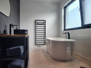 Une salle de bain moderne avec une baignoire, une étagère noire où sont posées des serviettes et une fenêtre qui laisse entrer la lumière naturelle.