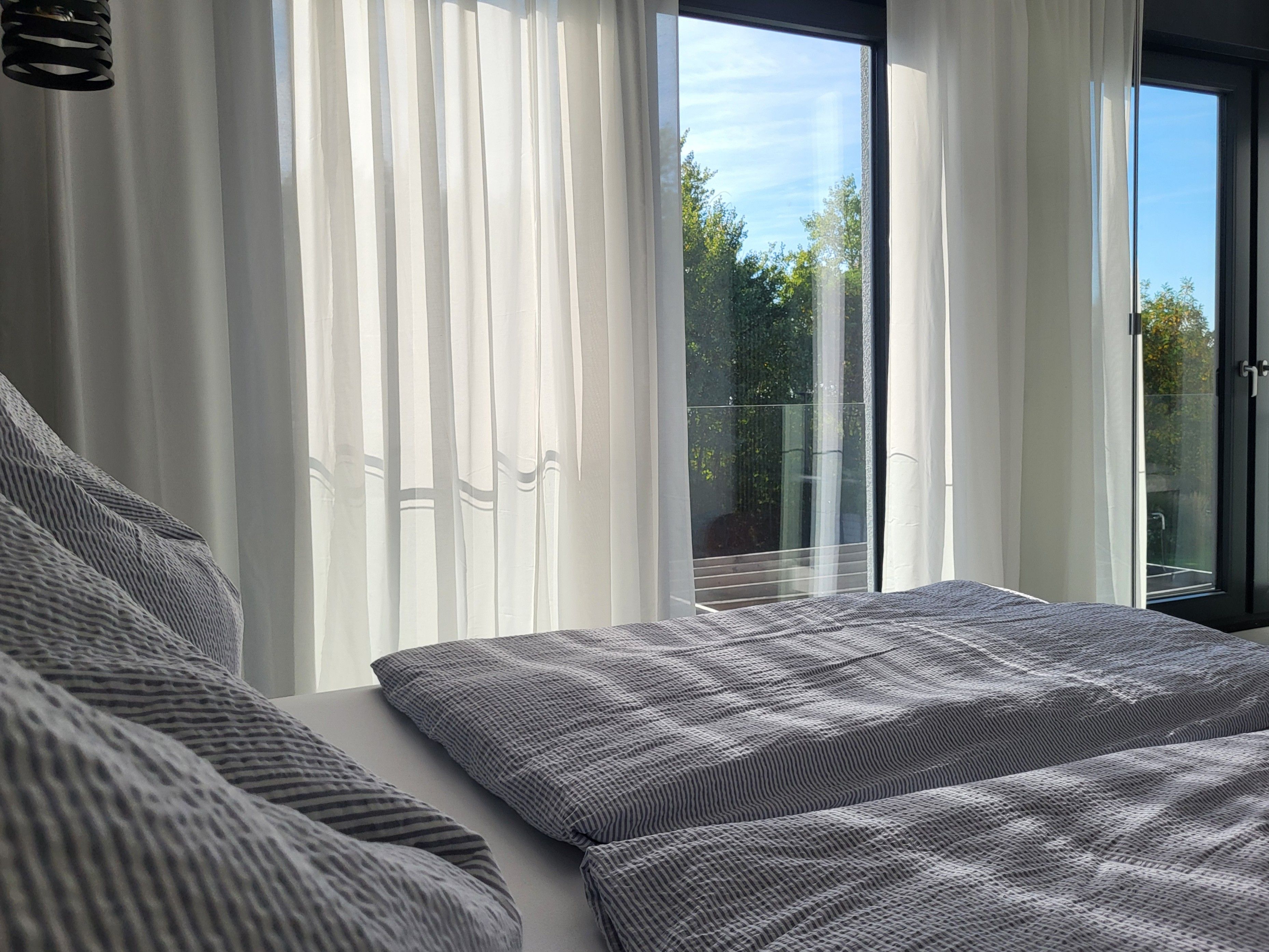 A bedroom with a large bed, white curtains, and a view of the outdoors through a glass door.