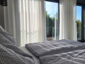 Une chambre avec un grand lit, des rideaux blancs et une vue sur l’extérieur grâce à une porte vitrée.