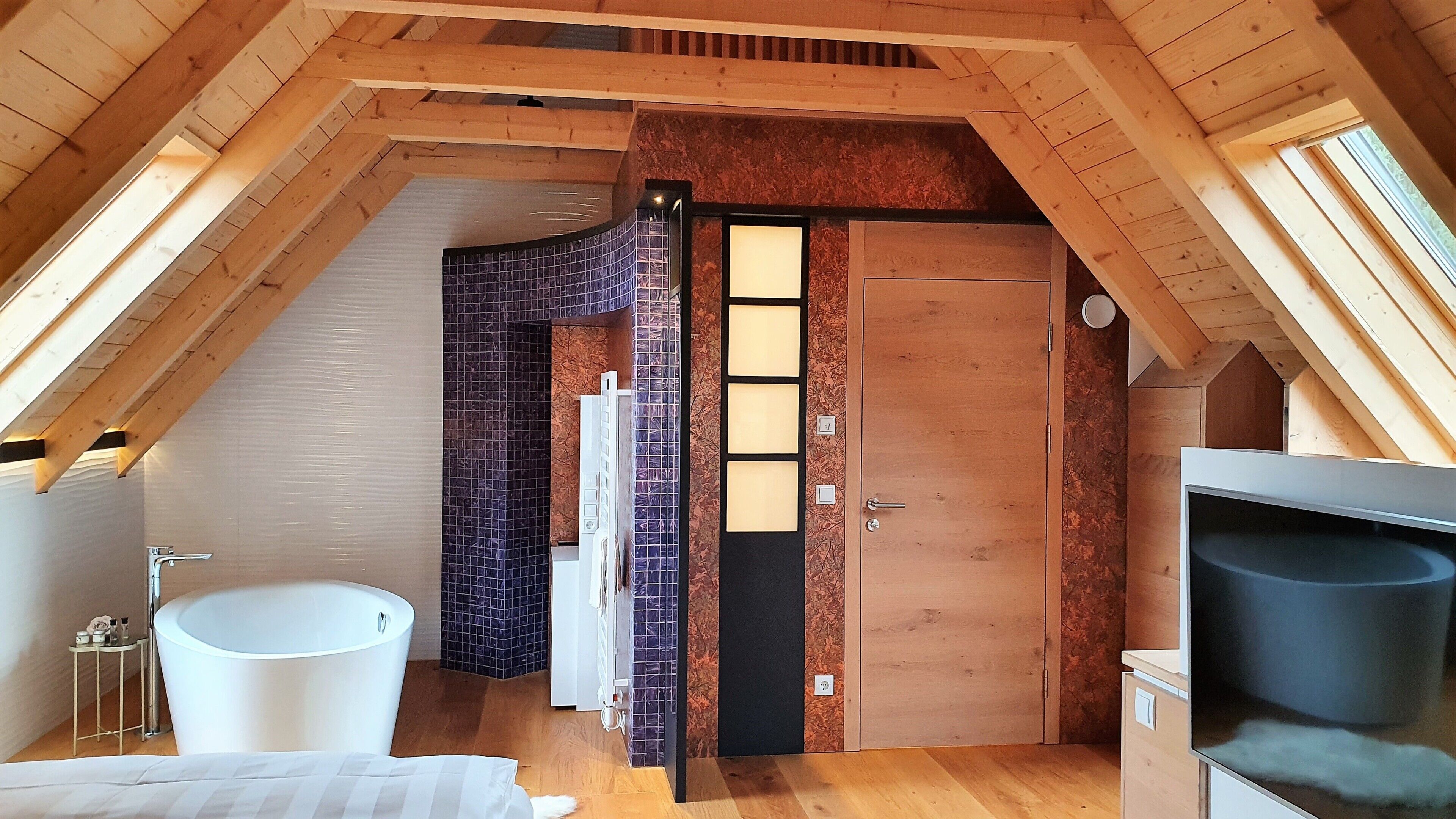 Ein Zimmer mit schrägen Wänden, einer Holztür, einer weißen Badewanne und einem kleinen Tisch mit Pflegeprodukten.