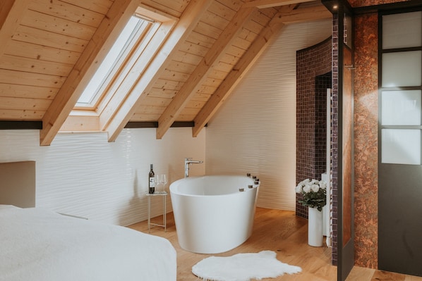 A modern bathroom with a freestanding bathtub, a small table with a bottle and glass, and a white rug on the floor.