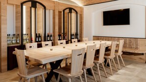 A long wooden table with chairs, a TV mounted on the wall, and wine bottles on shelves.