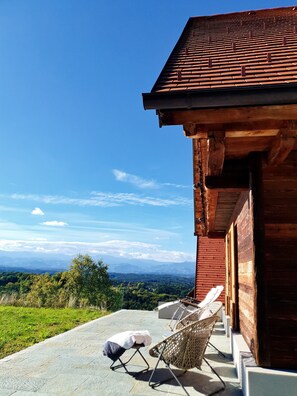 Un edificio de madera con techo de tejas, un área de estar exterior cubierta con sillas y vistas a montañas distantes.