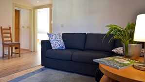 A living room with a dark grey sofa, a wooden chair, a small table with a lamp, and a potted plant.