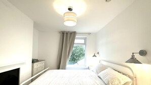 A bedroom with a bed, bedside table, lamp, and a window with curtains. - Modern & Brand New Apartment in Whitstable Centre (Whitstable)