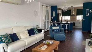 Un salon moderne avec un canapé blanc, des fauteuils bleus et une table basse en bois. On y trouve également un coin cuisine avec des tabourets de bar et un mur d’accent bleu.