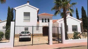 Une maison blanche à deux étages, avec un toit en tuiles, une allée bordée d’une grille et une voiture garée.
