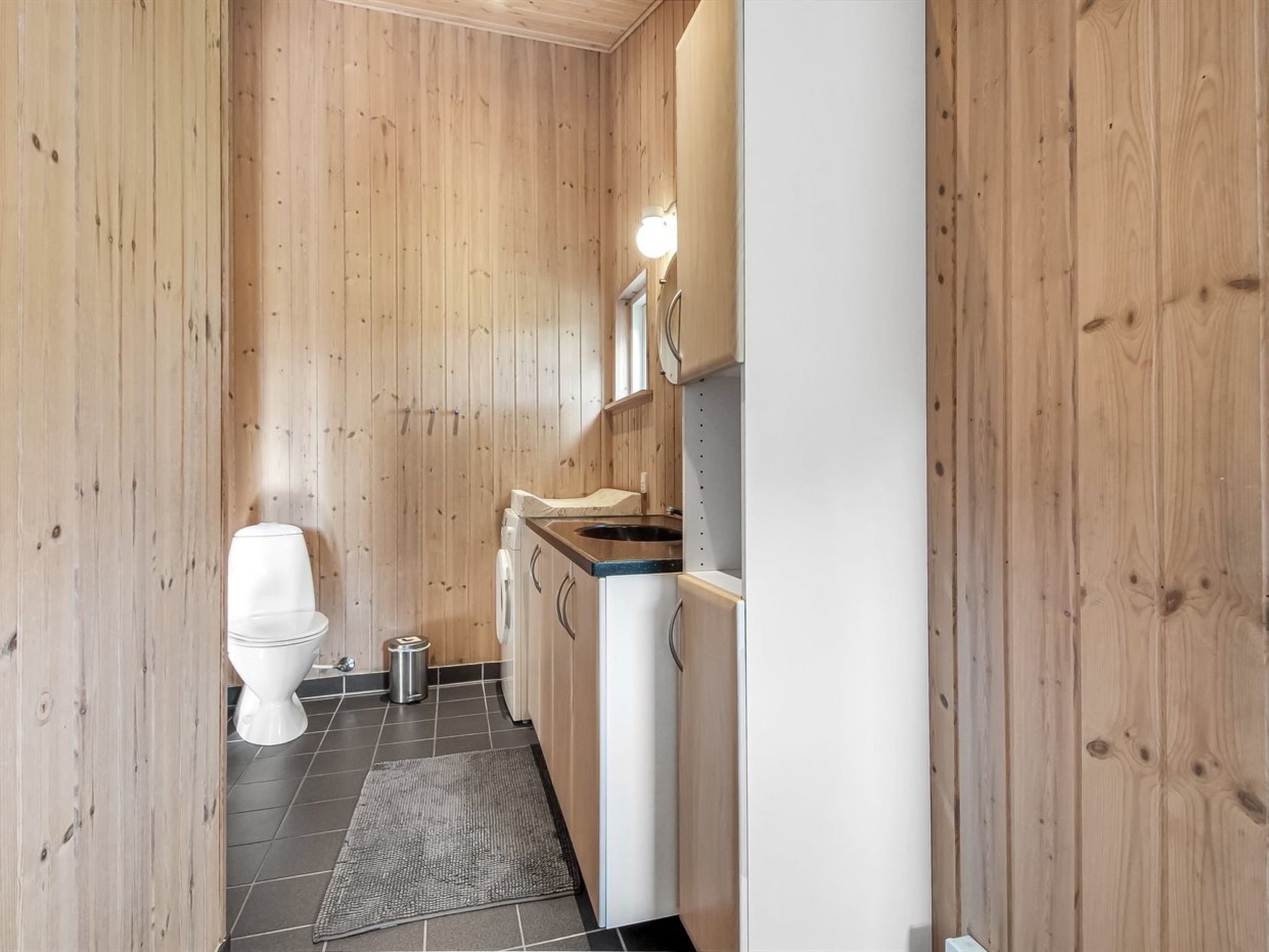 Ett litet badrum med träpanel på väggarna, en toalett, en tvättmaskin och ett handfat.