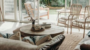 A cozy living room with wicker furniture, a round coffee table with decorative items, and a view of a garden through large glass doors. - Gypsea at the Bay, 26b Achilles St - Wi-Fi , air con, luxury and linen (Nelson Bay)