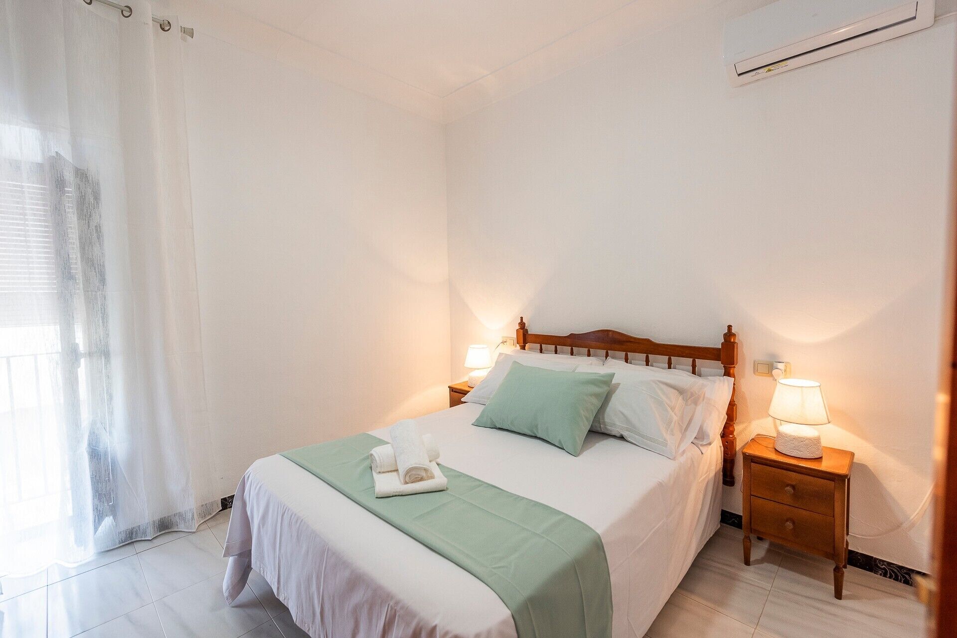Un lit bien fait, recouvert d’une couverture verte, avec deux oreillers et une serviette blanche. On y trouve deux tables de chevet avec des lampes, une fenêtre avec un rideau léger et un appareil de climatisation fixé au mur.