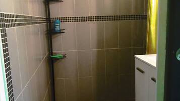 A small bathroom with a shower, a toilet, and a sink. There are shelves with toiletries and a green trash bin.