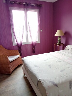 Une chambre à coucher avec un lit, une chaise, une fenêtre avec des rideaux et une table de chevet avec une lampe.