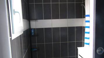 Ein kleines Badezimmer mit dunklen Fliesen, einer Dusche, einem weißen Waschbecken und einem blauen Mülleimer.