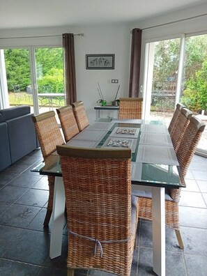 Un coin repas avec une table en verre entourée de chaises en osier, d’un canapé et une vue sur la verdure grâce à de grandes fenêtres.