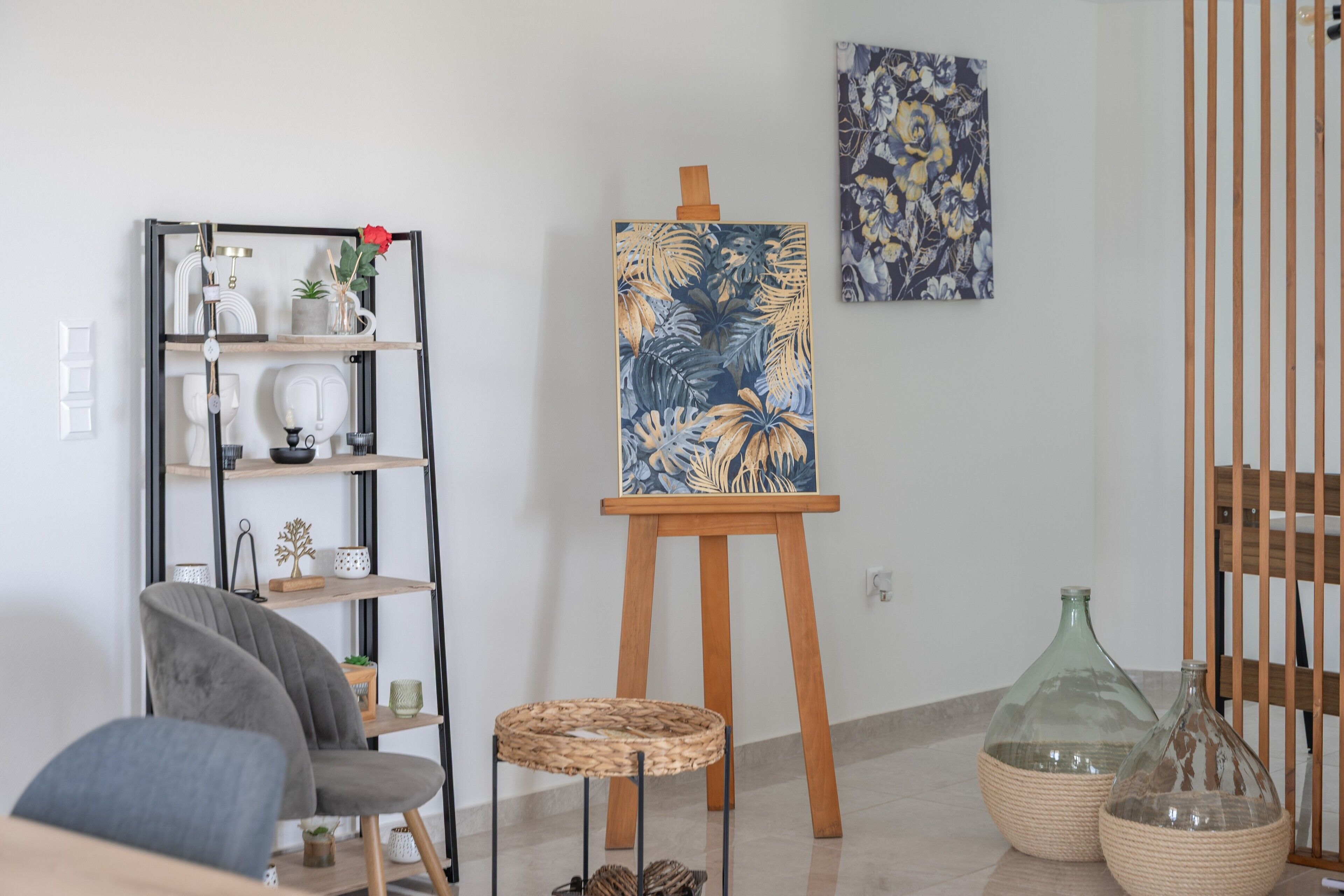 Ein Zimmer mit Staffelei, auf der eine Malerei steht, einem grauen Stuhl, einem gewebten Hocker und einem Regal mit Dekorationen.