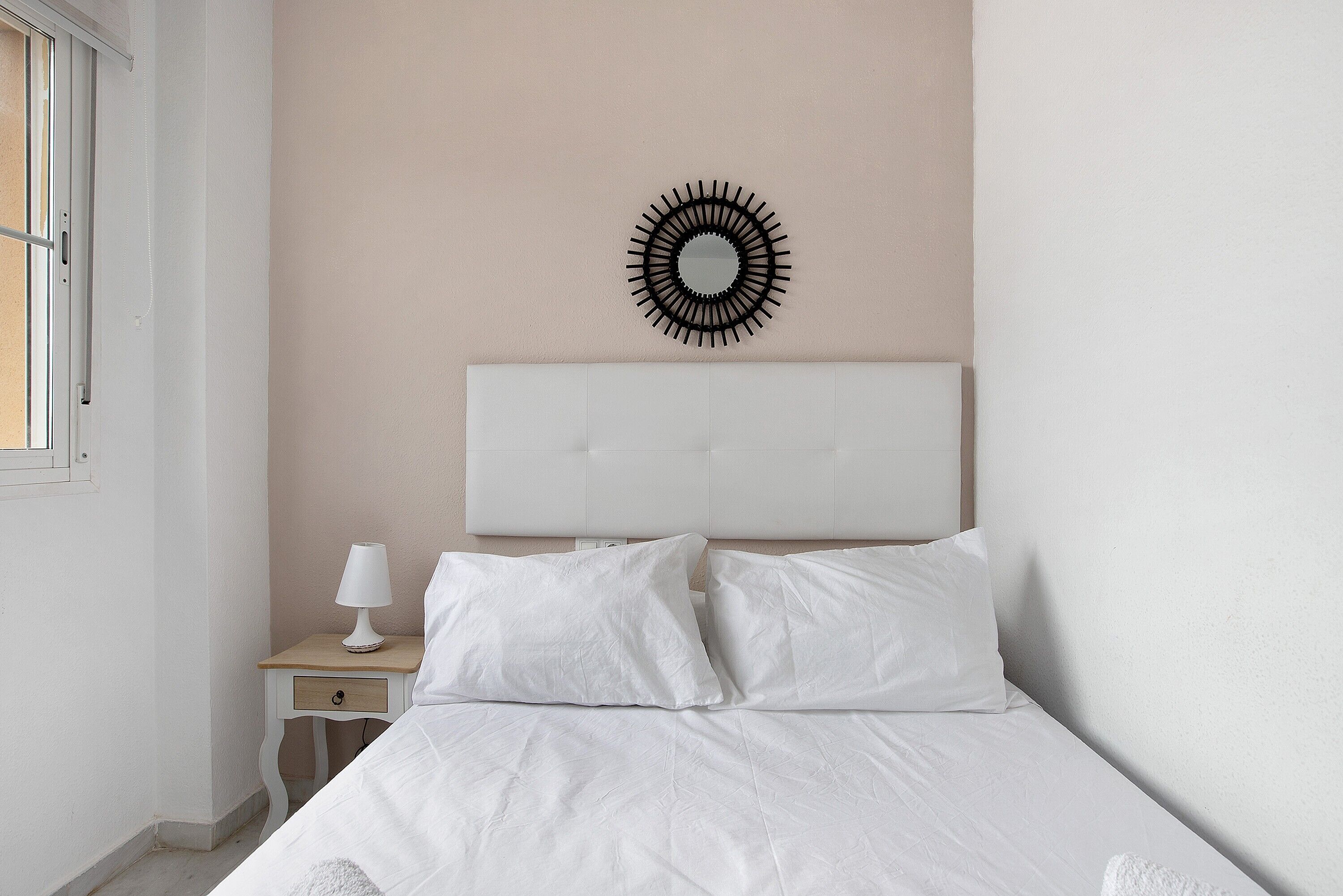 Une chambre à coucher avec un lit, une table de chevet avec une lampe, un miroir fixé au mur et une fenêtre.