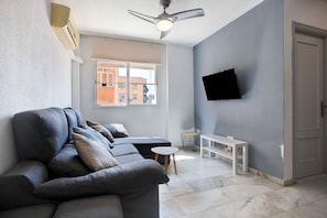 A living room with a sofa, a ceiling fan, a TV mounted on the wall, and a window with a view of buildings.