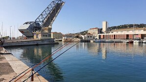 A waterfront with a large industrial structure, calm water, and buildings in the background. - F4 Crossing Sea View with Garages and Balconies (Sète)