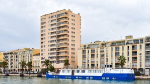 A tall residential building with balconies, a blue and white boat in the foreground, and palm trees along the waterfront. - F4 Crossing Sea View with Garages and Balconies (Sète)