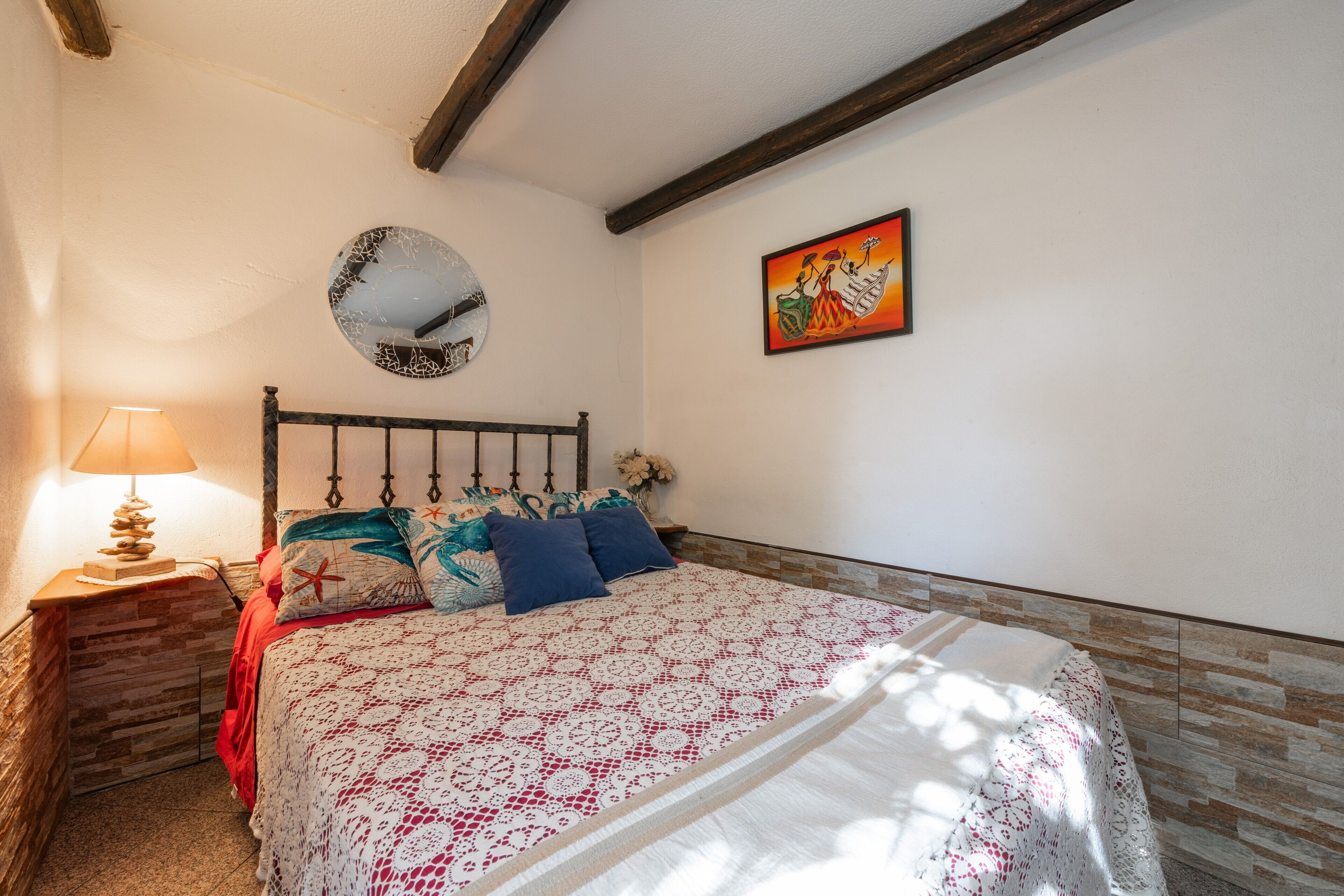Een slaapkamer met een bed, nachtkastje, lamp, ronde spiegel en een schilderij aan de muur.