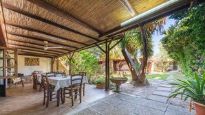 Una veranda esterna con tavoli e sedie in legno, un tetto di paglia e vista su un giardino.