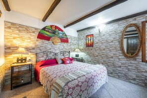A bedroom with a bed, two bedside tables, a wall-mounted fan, a decorative mirror, and a stone-patterned wall.