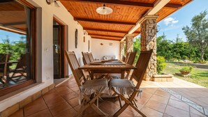 Um pátio externo com mesa e cadeiras de madeira, um pilar de pedra e vista para o jardim.
