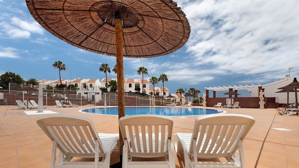 Área de piscina com cadeiras de sol brancas, um grande guarda-sol e uma piscina.