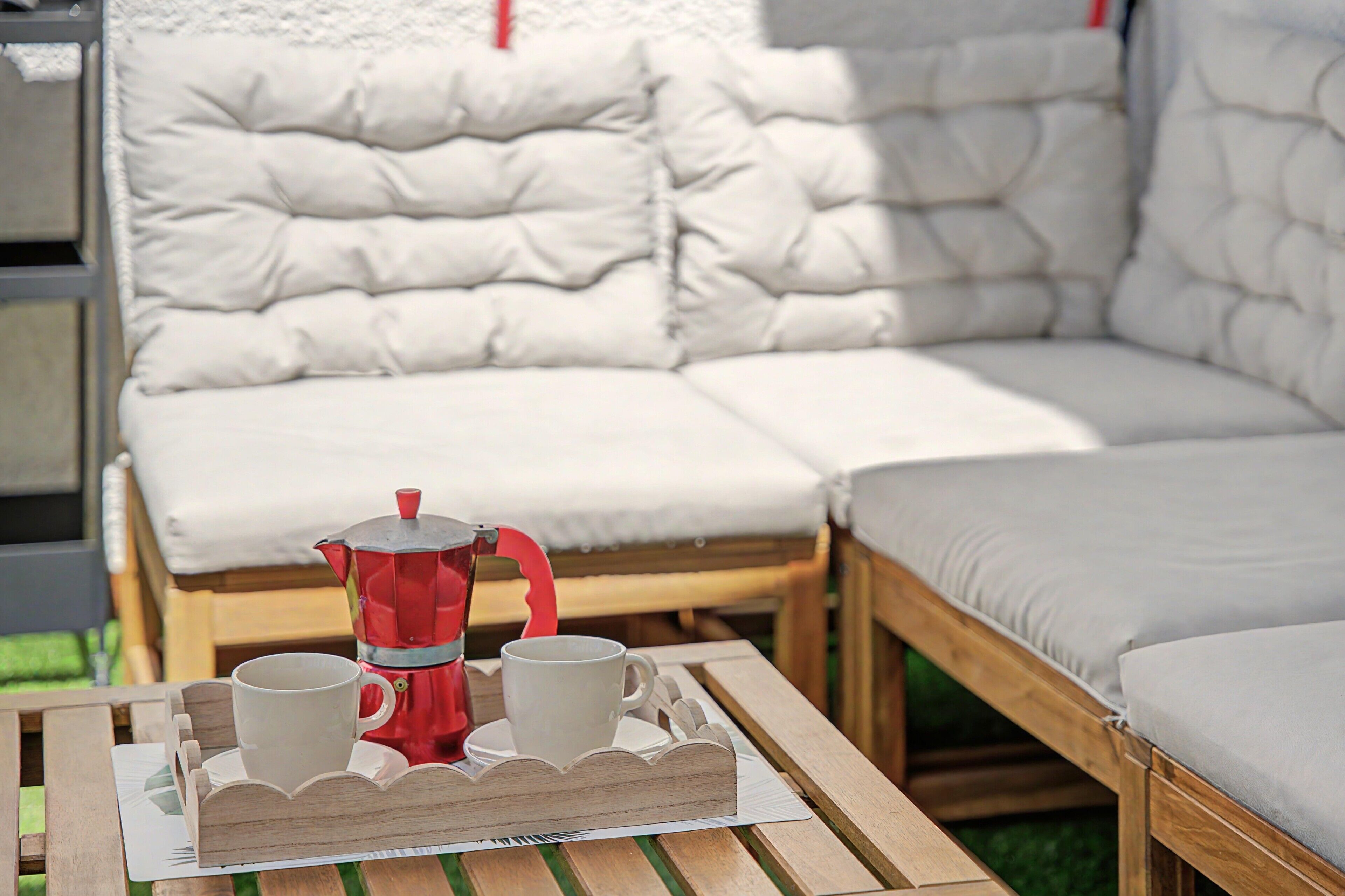 A wooden table with a tray holding a red coffee pot and two white cups, set on a patio with a cushioned outdoor seating area.