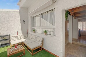 Ein gemütlicher Außenbereich mit einer Couch und einem Holztisch, ein Fenster mit Vorhängen und Blick ins Innere.