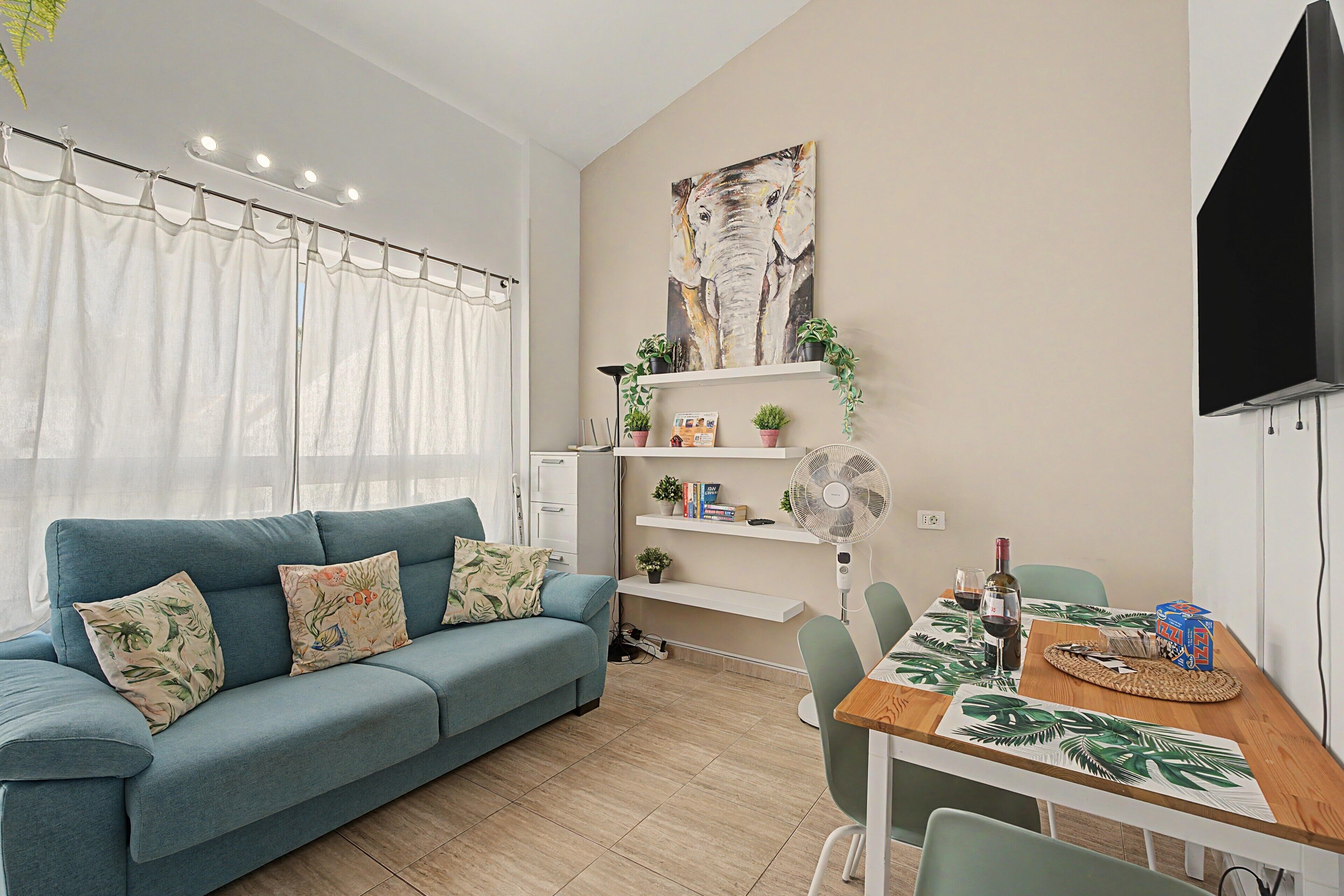 A modern living room with a blue sofa, a dining table with a tropical leaf table runner, and a mounted TV.