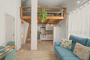 Un intérieur moderne avec un plafond de style industriel, une cuisine dotée de meubles blancs, un canapé bleu agrémenté de coussins à motifs et un coin repas avec une chaise verte.
