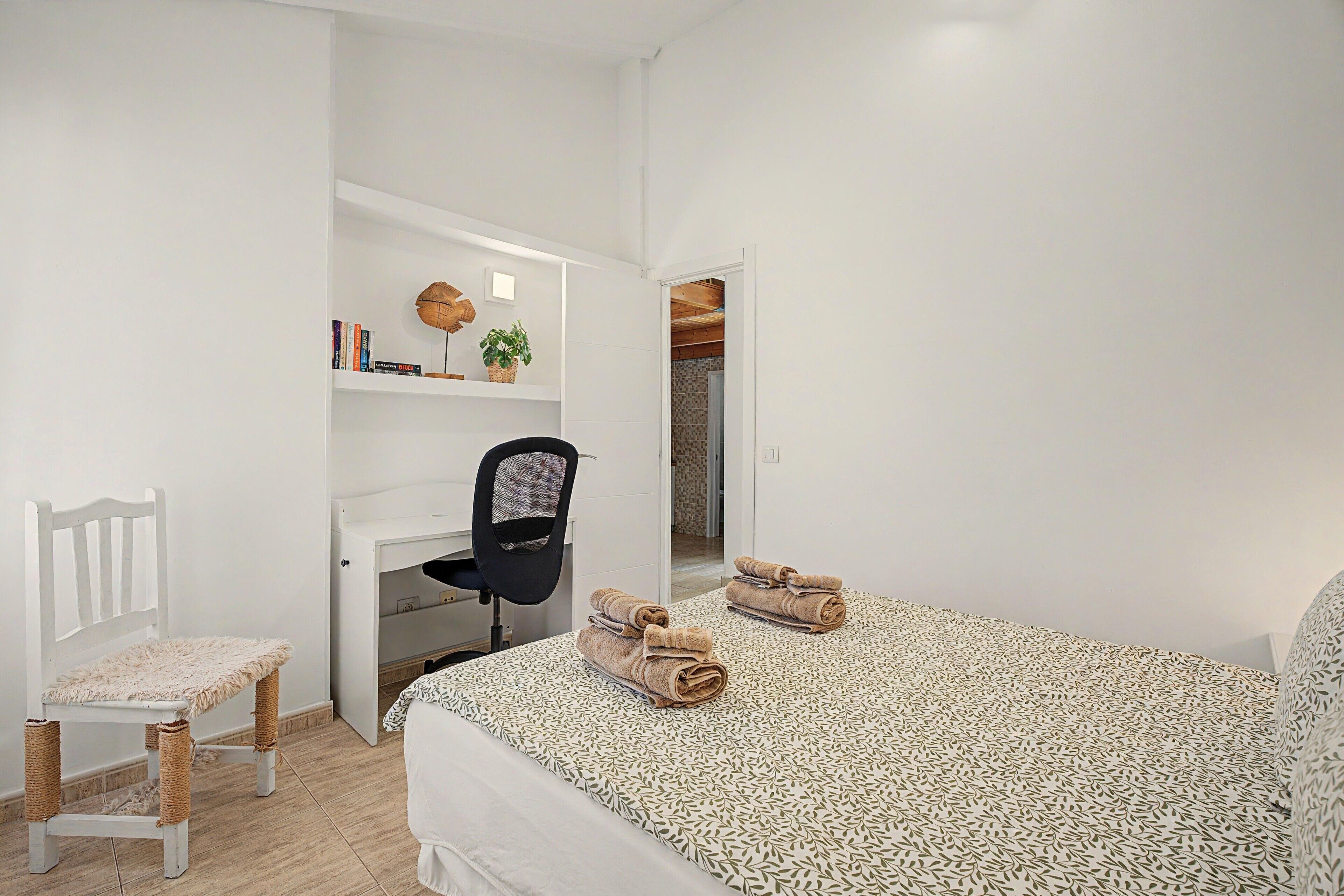 A bedroom with a bed, a desk, a chair, and a small table with books and a plant.